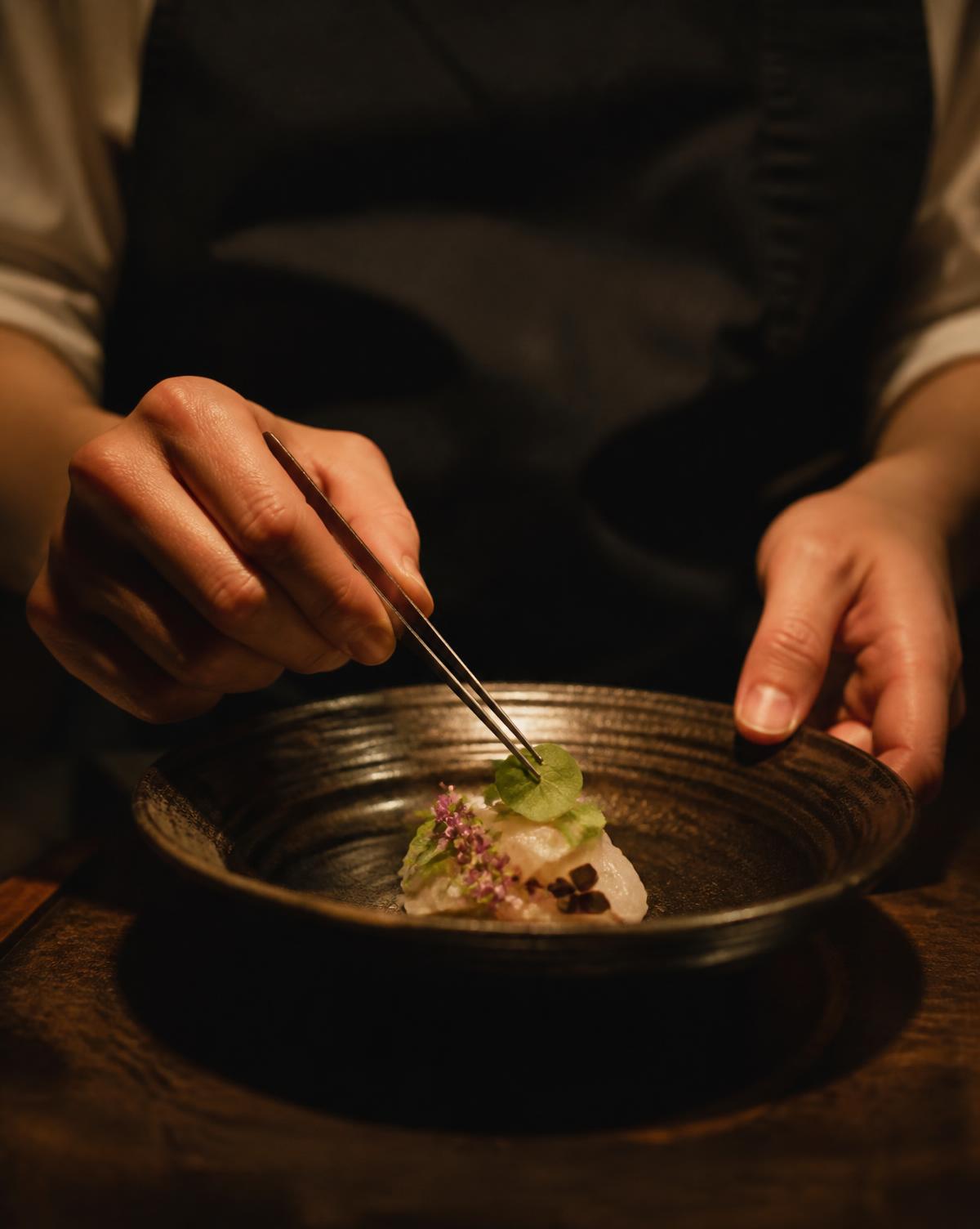 Chef plating a dish with tweezers under warm kitchen light